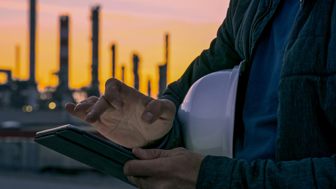 Close-up shot of the hands an engineer with a white hardhat using a tablet with an oil refinery visible in the background during sunset